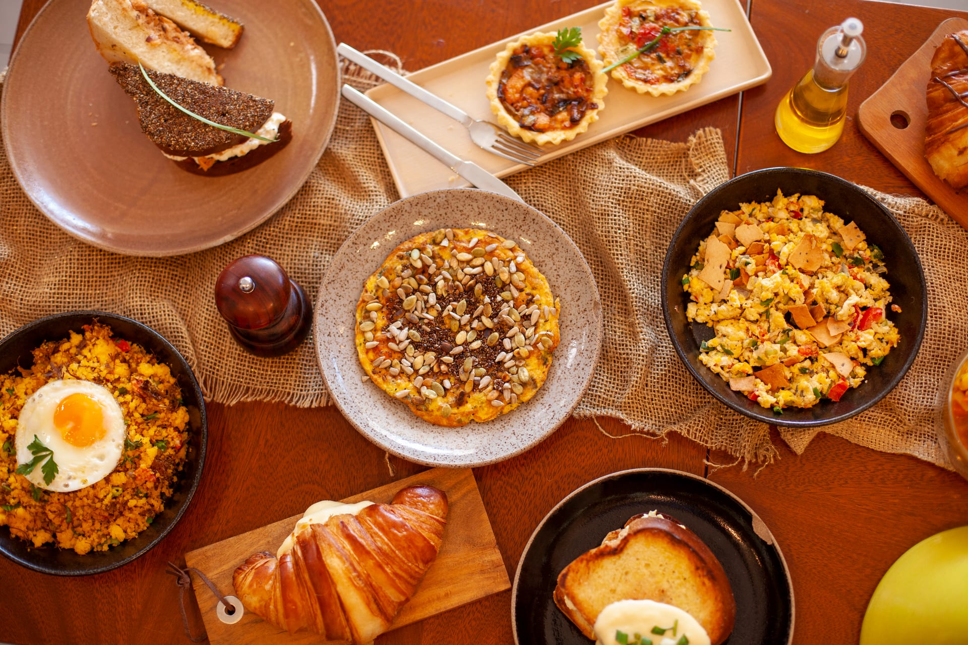 Mesa farta com variedade de pratos: cuscuz com ovo, croissant, quiches, ovos mexidos, pão na chapa, tapioca com sementes